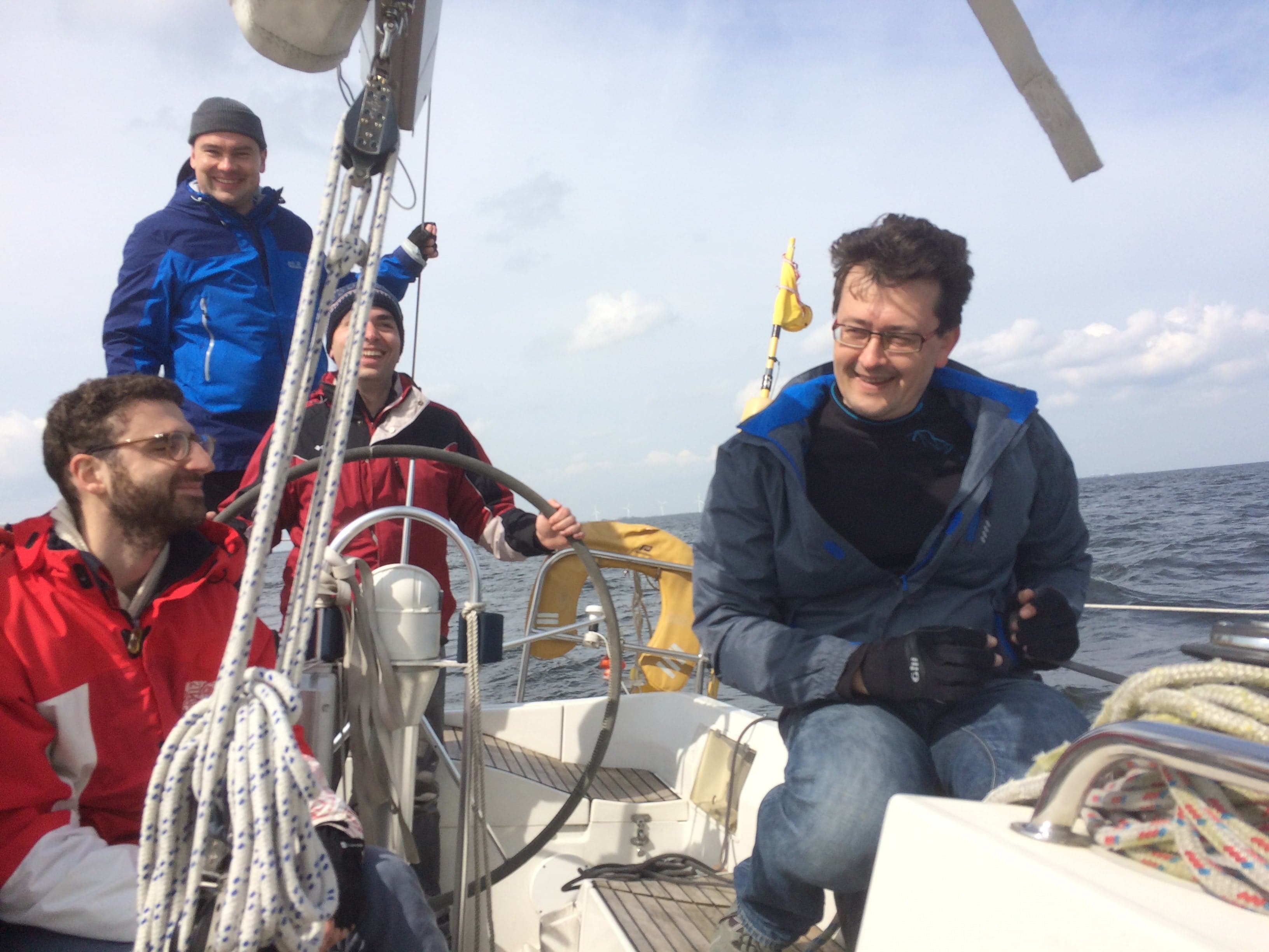 Four smiling crew members relaxing in the cockpit of a yacht, wind turbines on the horizon.